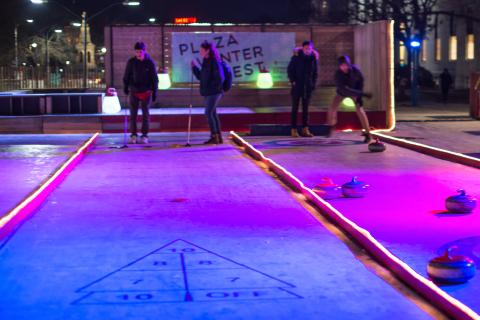 Photograph of people playing shuffleboard and curling on synthetic ice lanes on the Science Center Plaza at night with glowing lights 
