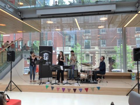 Photo of the stage in Harvard Commons with a singer, drummer, guitarist, bassist, and Club Passim banner