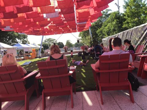photo of people sitting on adirondak chairs beneath the plaza's shade canopy with performer in the distance